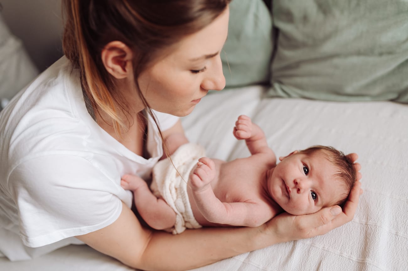 Mama z noworodkiem w sypialni – ciepła, rodzinna atmosfera domowej sesji fotograficznej.