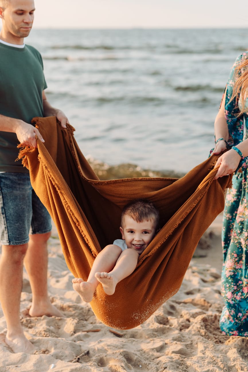 Roześmiany chłopiec huśtany w kocu w trakcie nadmorskiej sesji o zachodzie słońca. 