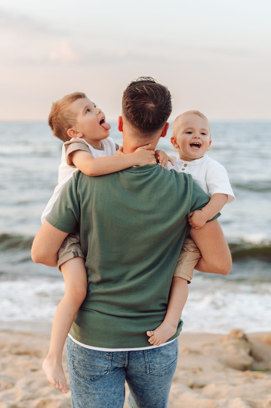 Tata spogląda na morze trzymając synków, podczas gdy jeden z nich próbuje go polizać.